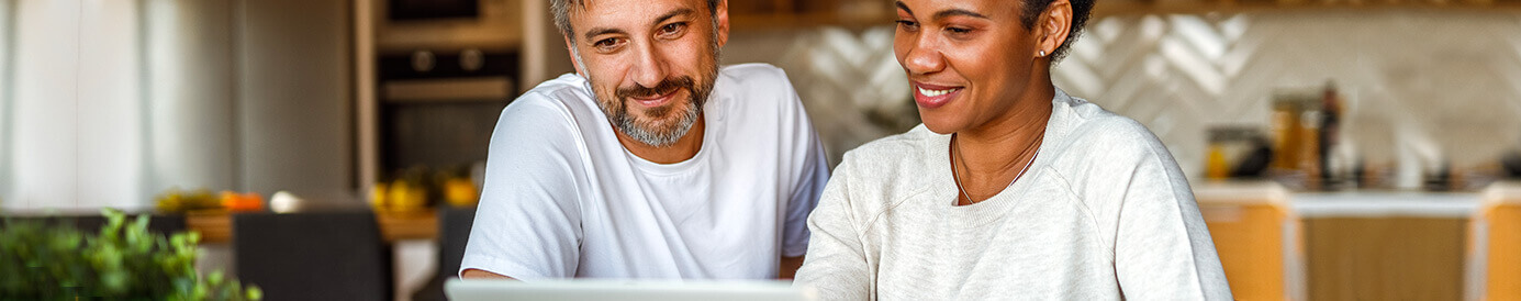A couple sits at the kitchen counter while looking at a laptop together