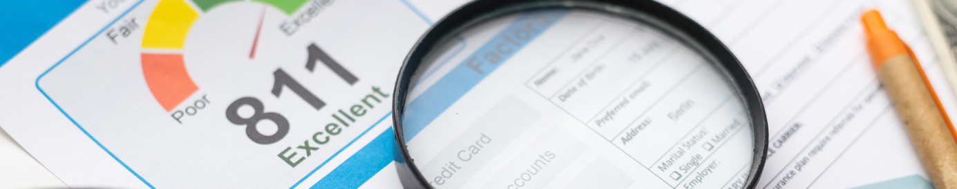 A magnifying glass rests on a surface with documents related to credit. A pen, glasses and a 100 dollar bill lay beside it.