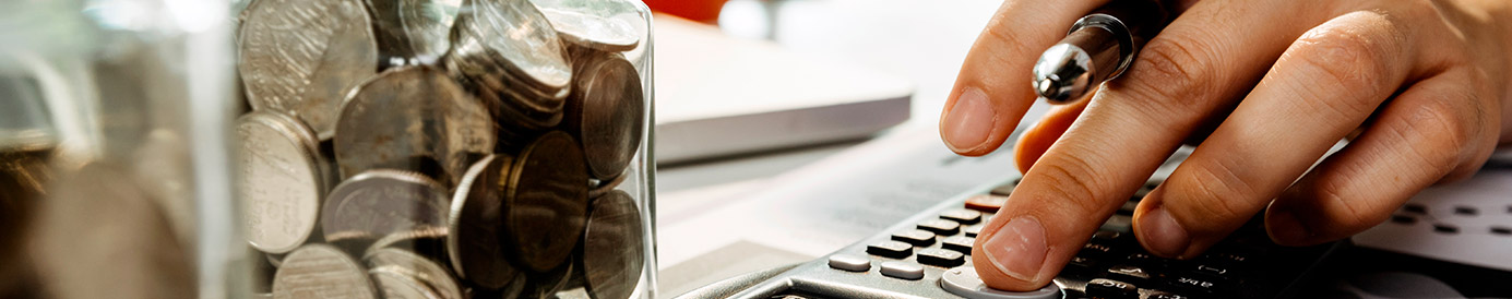 A person is reading from their iPad while typing into a calculator. There is a coffee mug and some piggy banks next to the calculator on the desk.