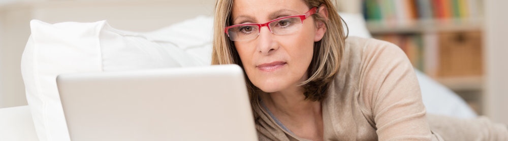 A woman wearing red glasses and a beige shirt is reading something on her laptop.