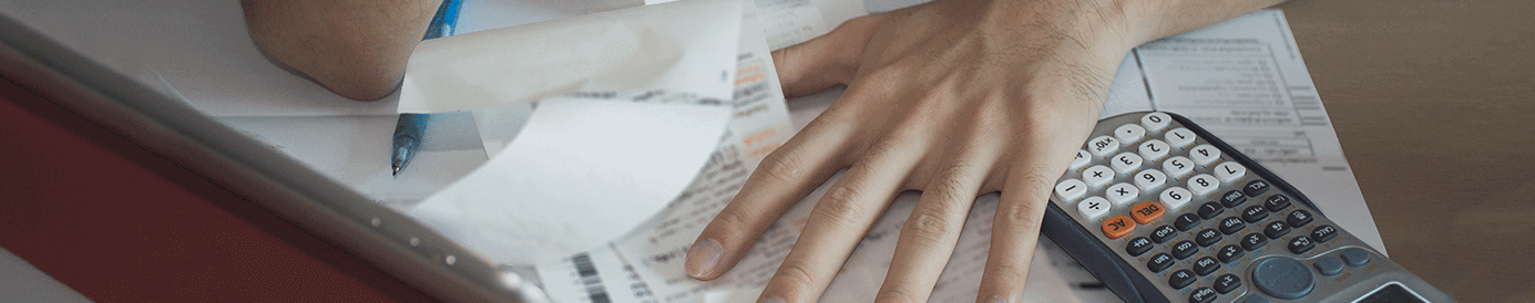 A man sits at a desk while looking at bills and receipts. He has a pen and calculator.